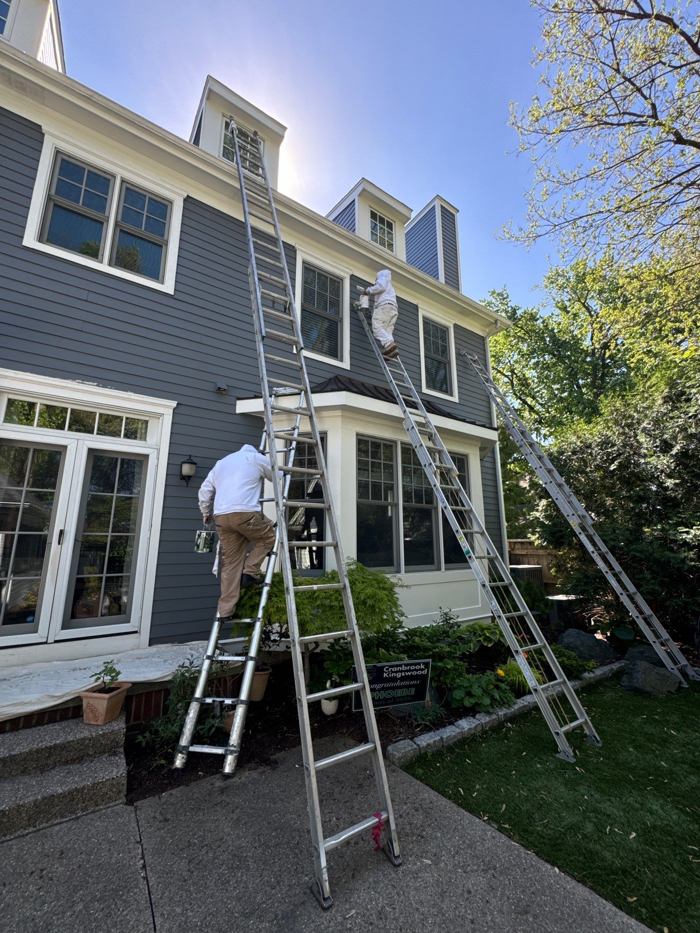 Experienced Exterior House Painters at Work Two professional painters on ladders applying fresh paint to the exterior of a gray house with white trim.