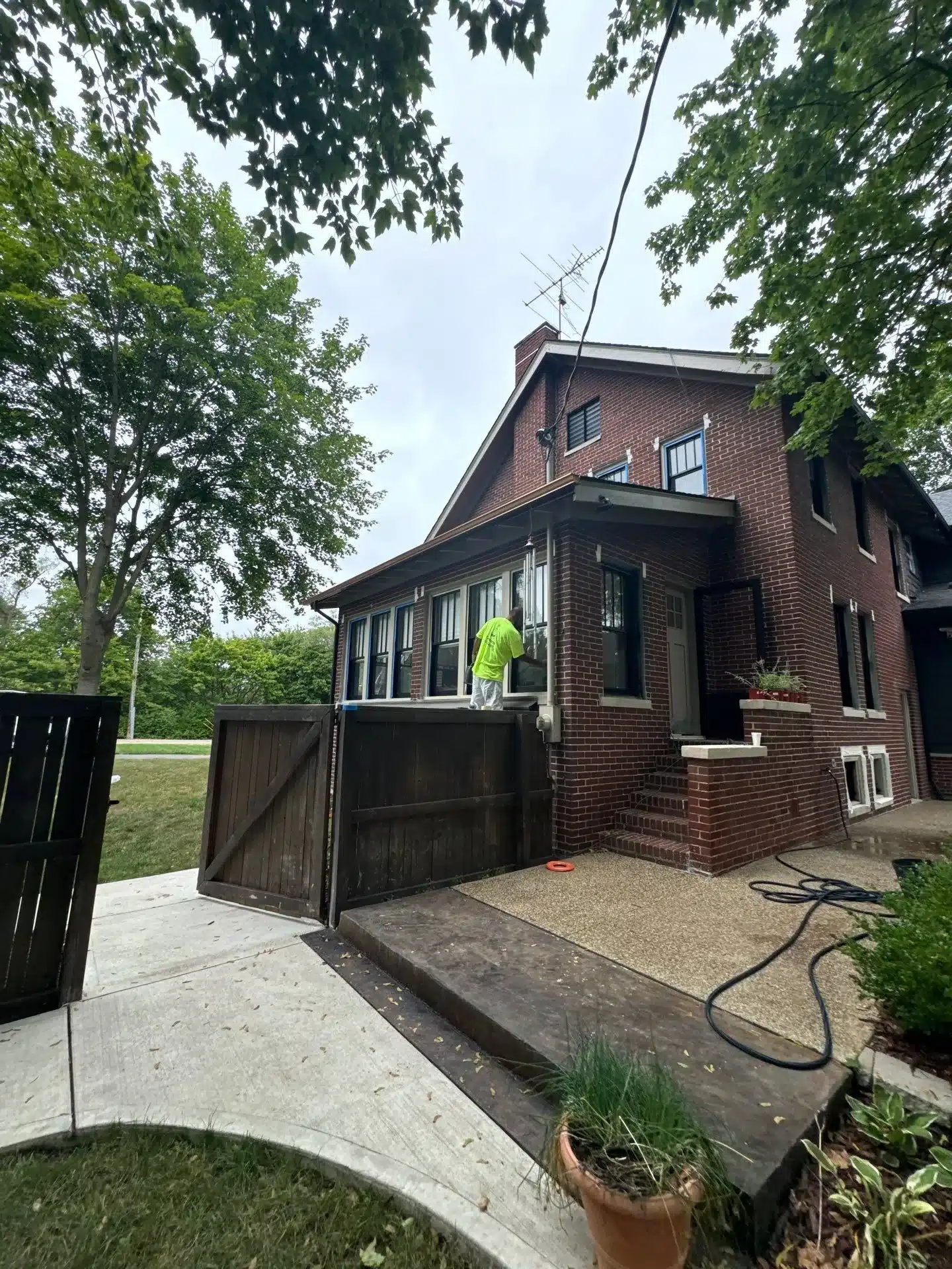 Brick Home Exterior Painting Project Red brick two-story residential home with dark-trimmed windows, painter on ladder, and dark wooden gate during exterior
